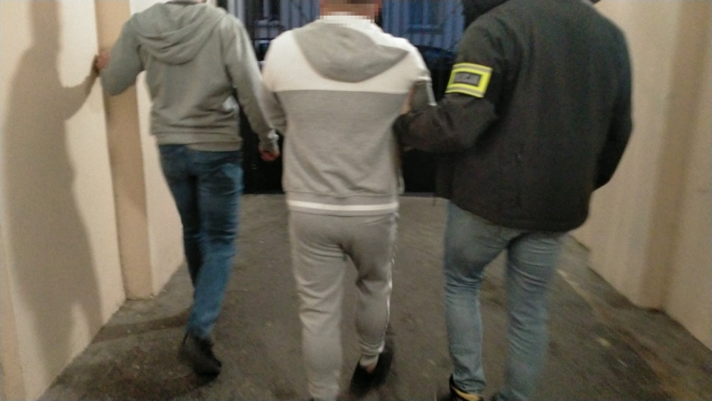 Trzech mężczyzn idzie korytarzem, jeden z nich w szarym dresie jest prowadzony przez funkcjonariusza w kurtce z oznaczeniem.