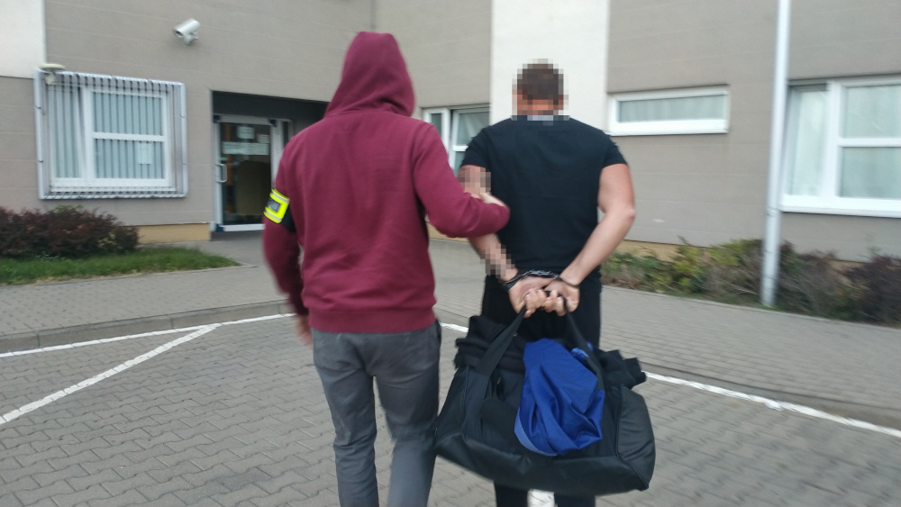 Funkcjonariusz w czerwonym kapturze prowadzi zatrzymanego mężczyznę w kajdankach, trzymającego czarną torbę.