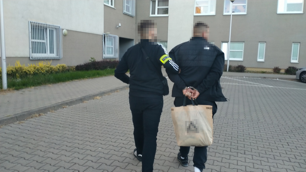Funkcjonariusz w mundurze prowadzi zatrzymanego mężczyznę z rękami związanymi na plecach w otoczeniu budynków mieszkalnych.