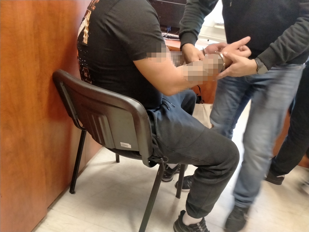 Mężczyzna w ciemnej koszulce siedzi na krześle, podczas gdy funkcjonariusze policji zakładają mu kajdanki w pomieszczeniu.