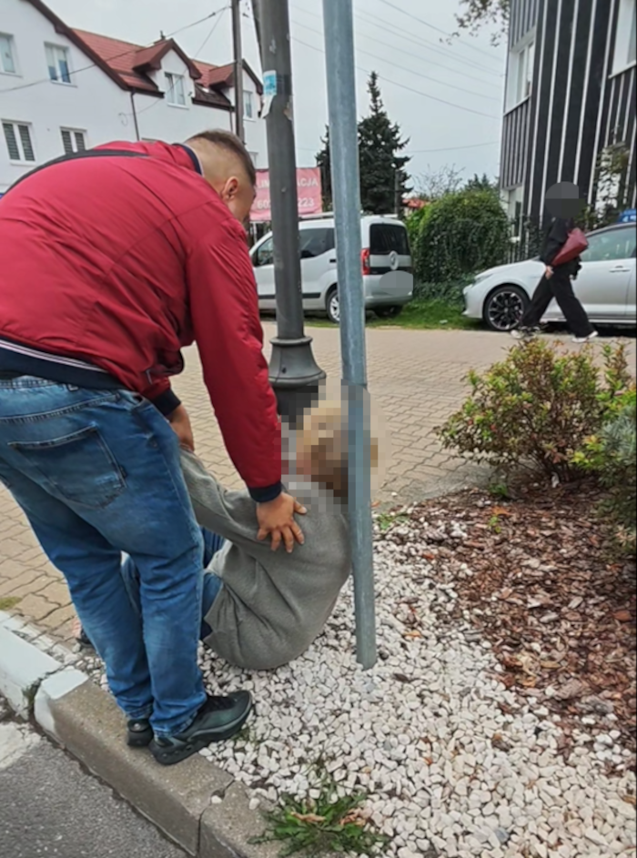 Mężczyzna w czerwonej kurtce pomaga osobie siedzącej na ziemi obok słupa, w miejskim otoczeniu z samochodami w tle.