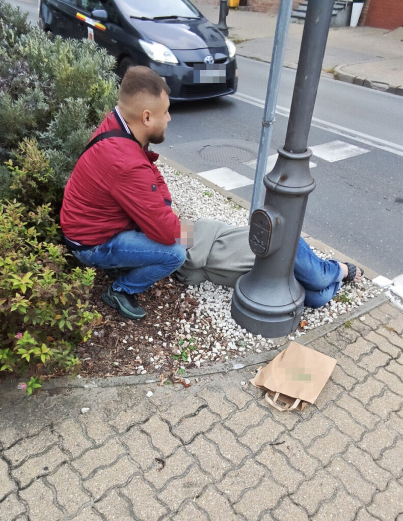 Mężczyzna w czerwonej kurtce siedzi obok osoby leżącej na trawniku przy latarni ulicznej.