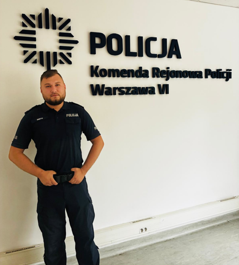 Mężczyzna w mundurze policjanta stoi w biurze przy ścianie z napisem "POLICJA Komenda Rejonowa Policji Warszawa VI".