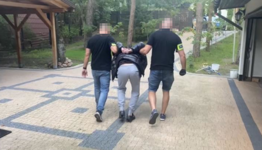 Mężczyzna w czarnej kurtce prowadzony przez dwóch funkcjonariuszy policji w otoczeniu zieleni i drewnianych budynków.