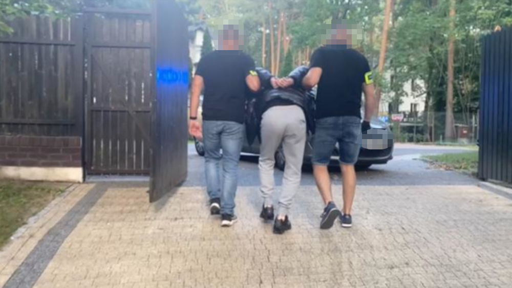 Mężczyzna w dresie prowadzony przez dwóch policjantów w mundurach w otoczeniu drzew i zabudowań.