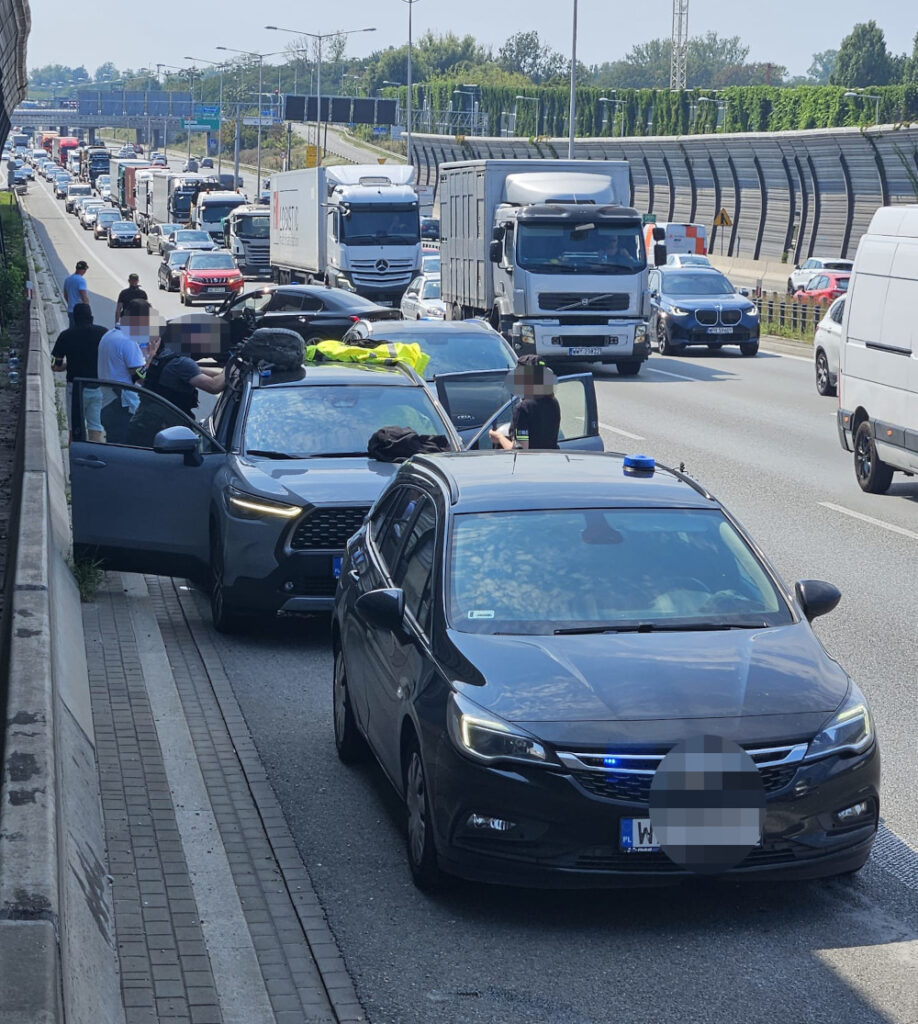 Grupa osób w kamizelkach odblaskowych przeszukuje pojazdy na zablokowanej drodze, obok stoją policyjne samochody.