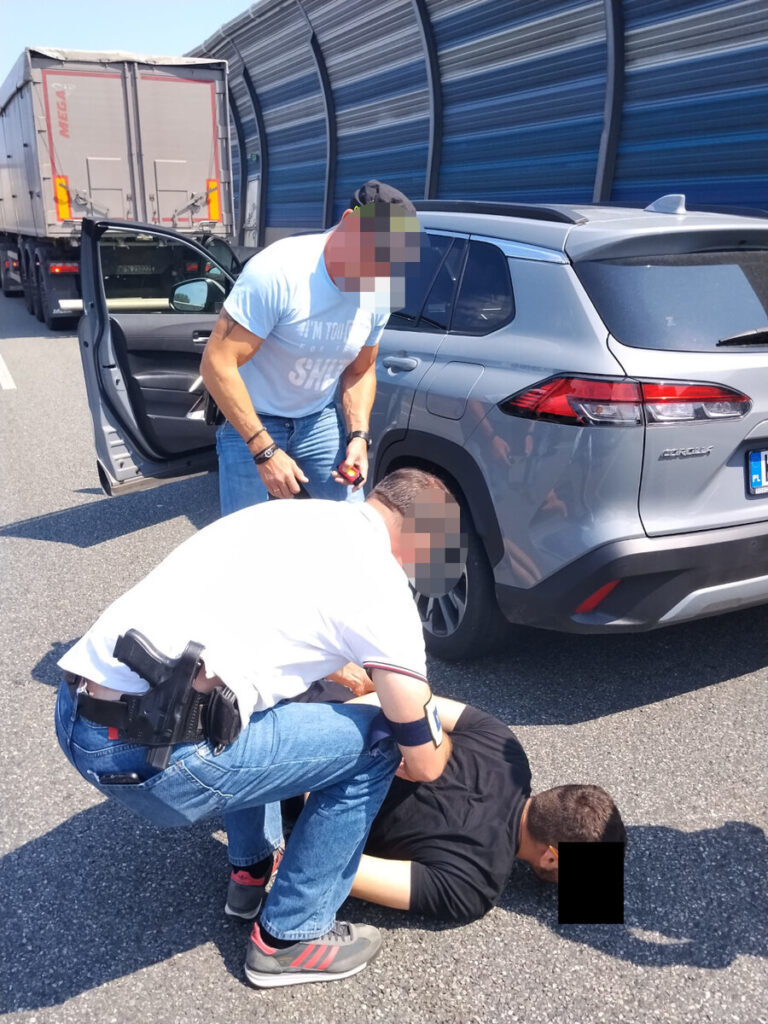 Mężczyzna w niebieskiej koszulce kontroluje leżącego na jezdni mężczyznę, podczas gdy drugi mężczyzna w białej koszuli go przytrzymuje. W tle widać zaparkowane samochody ciężarowe.