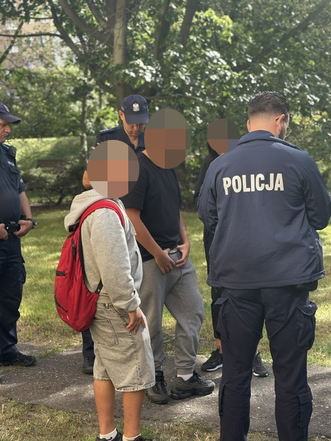 Grupa policjantów rozmawia z młodymi mężczyznami w parku, w tle widoczne drzewa.