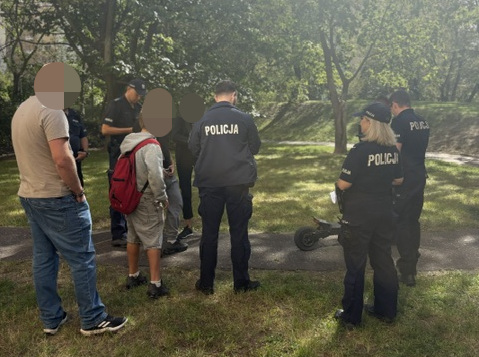 Grupa policjantów rozmawia z kilkoma osobami w parku, w tle drzewa i trawa.