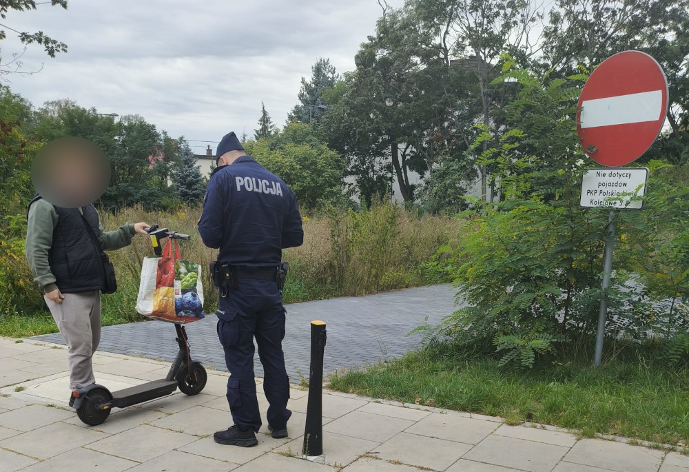 Mężczyzna na hulajnodze rozmawia z policjantem na chodniku, obok znaku zakazu wjazdu. W tle zarośla i budynki.