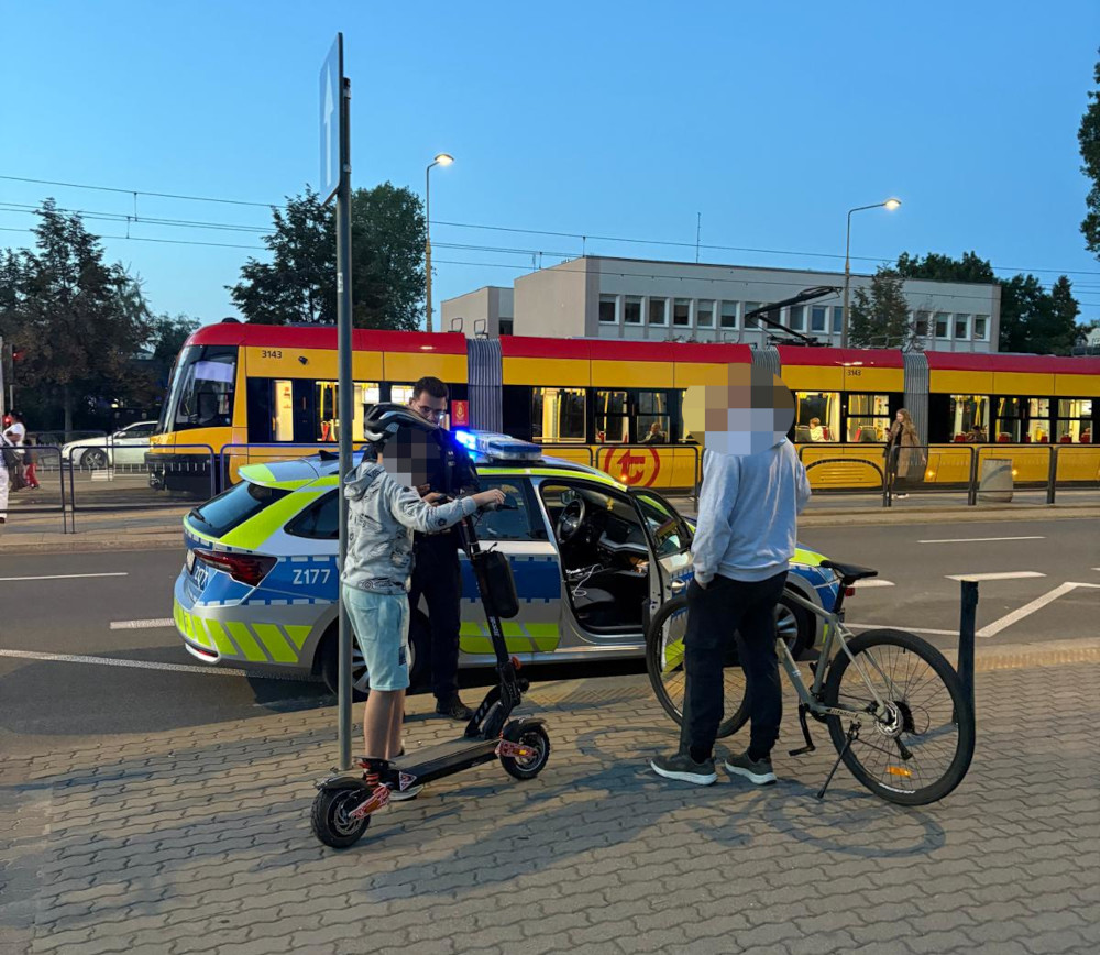Młody mężczyzna na hulajnodze elektrycznej rozmawia z policjantem obok radiowozu w miejskim otoczeniu.