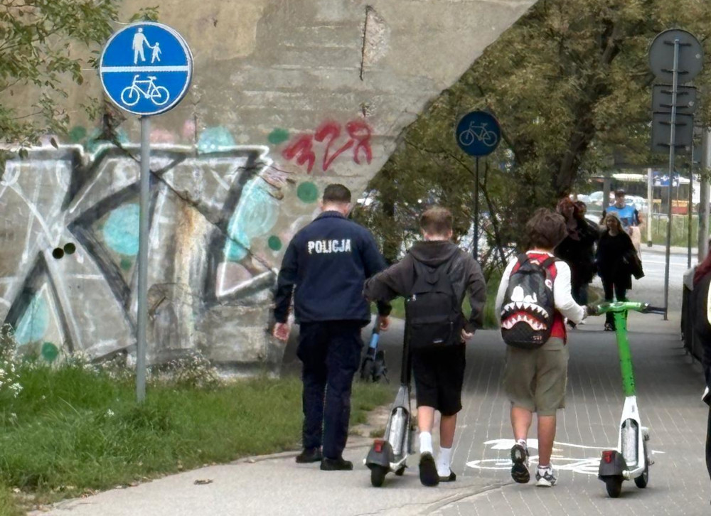 Policjant sprawdzający kierujących hulajnogami elektrycznymi na ścieżce rowerowej obok graffiti.