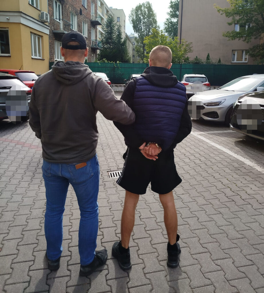 Mężczyzna w ciemnej bluzie trzyma rękę na ramieniu mężczyzny w kajdankach, stojących na parkingu.