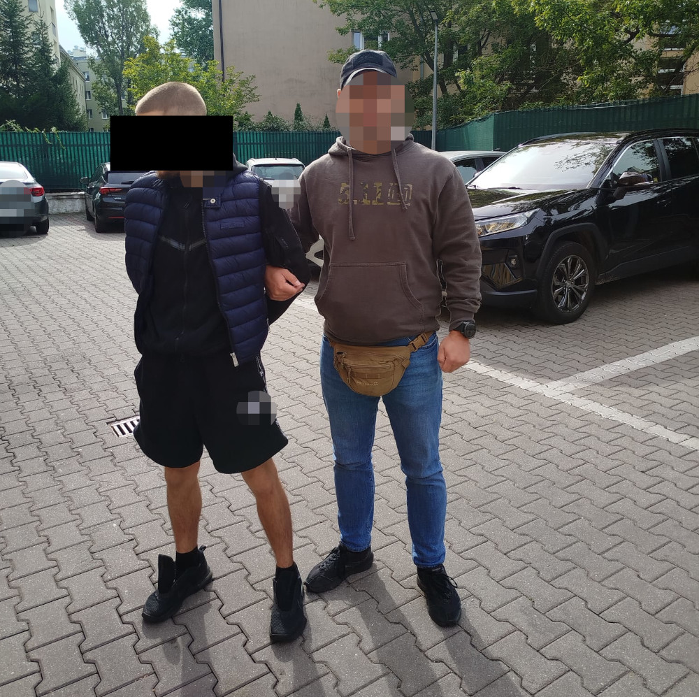 Mężczyzna w krótkich spodniach i kamizelce puchowej prowadzony przez osobę w brązowej bluzie i z torbą, na parkingu.