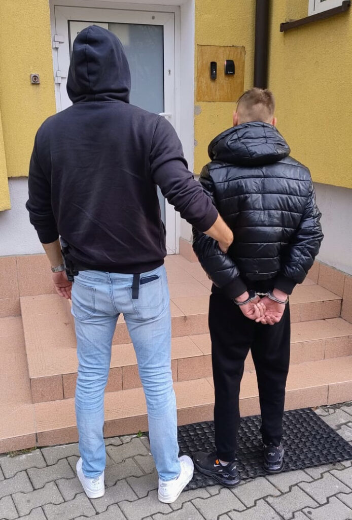 Mężczyzna w kapturze prowadzi skute kajdankami ręce mężczyznę w puchowej kurtce przed wejściem do budynku.
