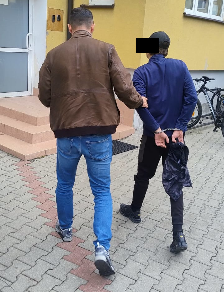 Mężczyzna prowadzi osobę z zakutymi z tyłu rękami w stronę wejścia do budynku z żółtą elewacją.