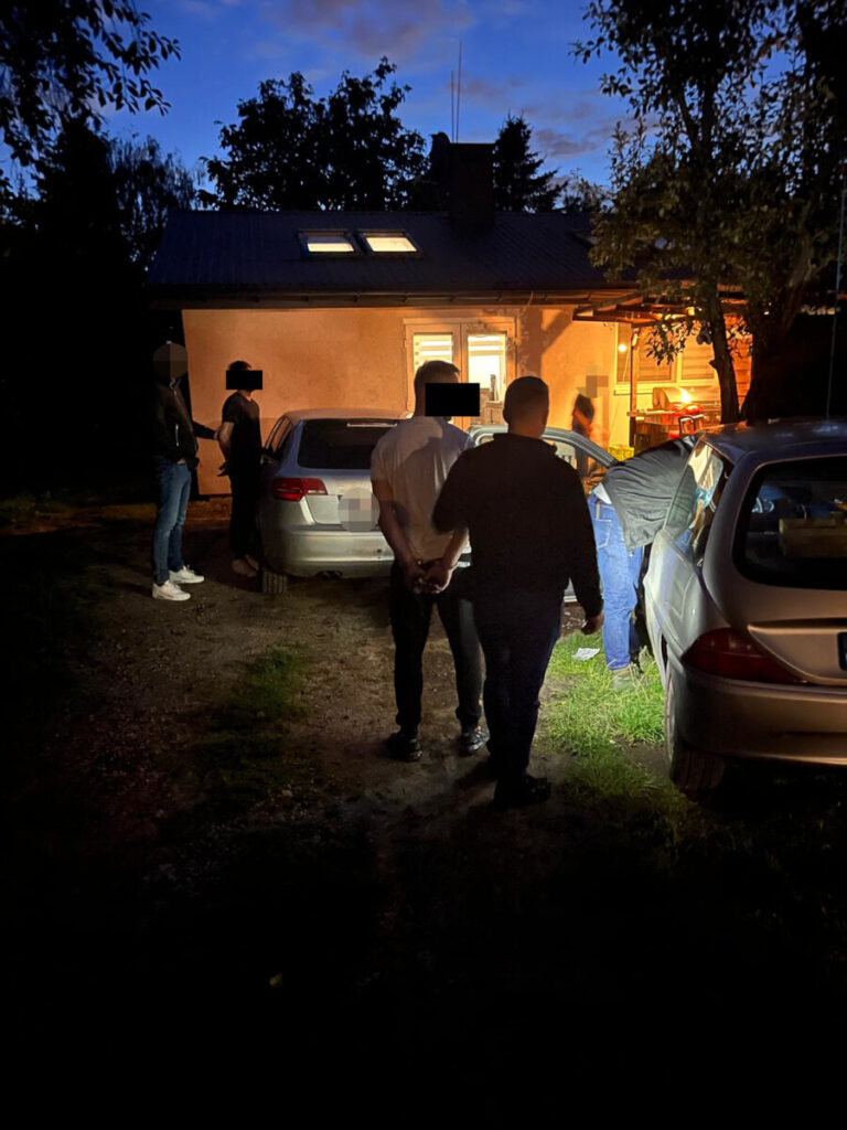 Mężczyzna z rękami za plecami prowadzony nocą między samochodami przy oświetlonym domu, obok stoją inni ludzie.