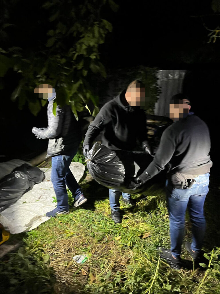 Trzej mężczyźni przenoszą czarne worki na zarośniętym terenie nocą.