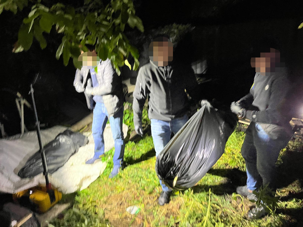 Trzech mężczyzn przenosi duży czarny worek w oświetlonym latarkami, zadrzewionym terenie nocą.