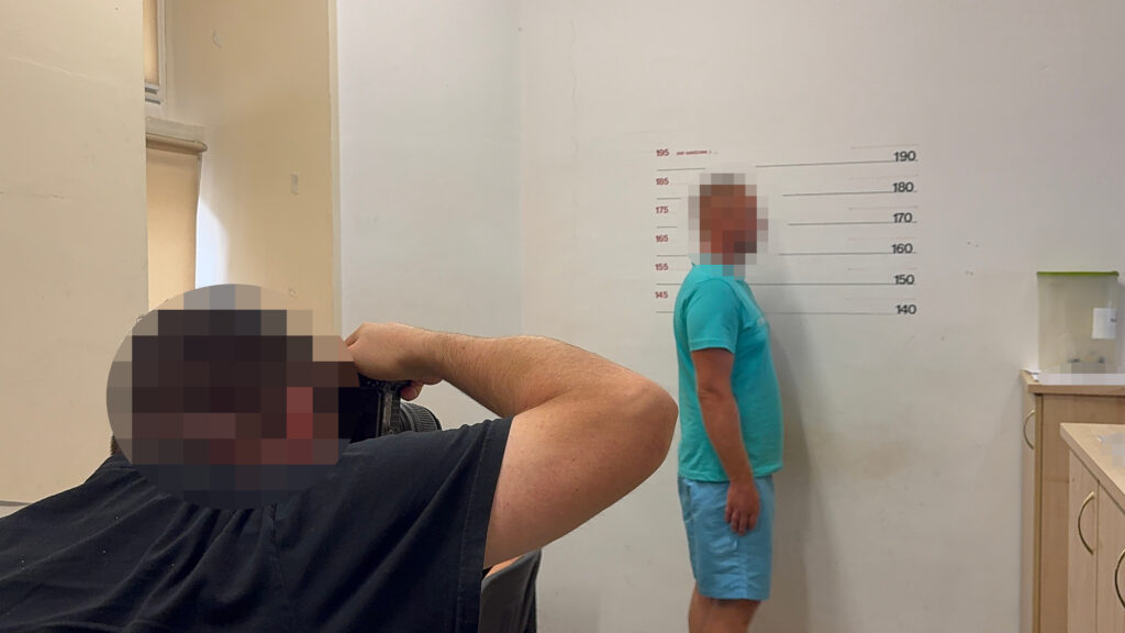 Mężczyzna w niebieskiej koszulce stoi przy ścianie z oznaczeniami wysokości, podczas gdy inny mężczyzna robi mu zdjęcie.
