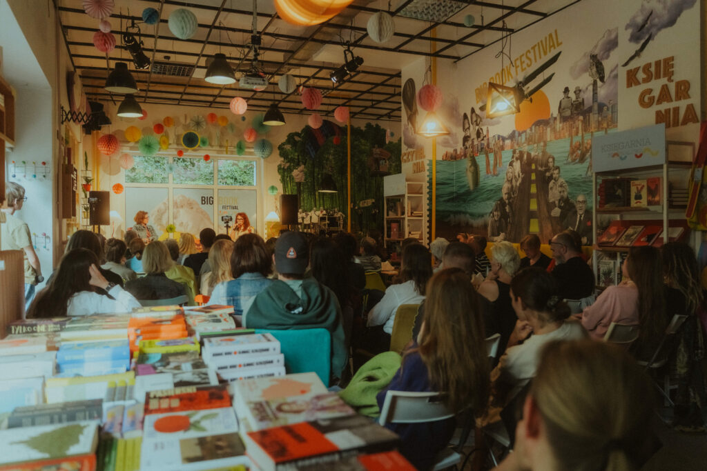 Tłum ludzi siedzi w kawiarni, słuchając dwóch prelegentek rozmawiających na scenie podczas festiwalu literackiego.