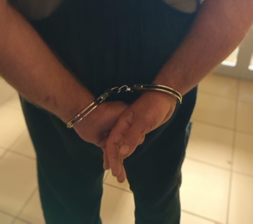 Mężczyzna z rękami skuty w kajdankach, stojący na jasnej podłodze w zamkniętym pomieszczeniu.