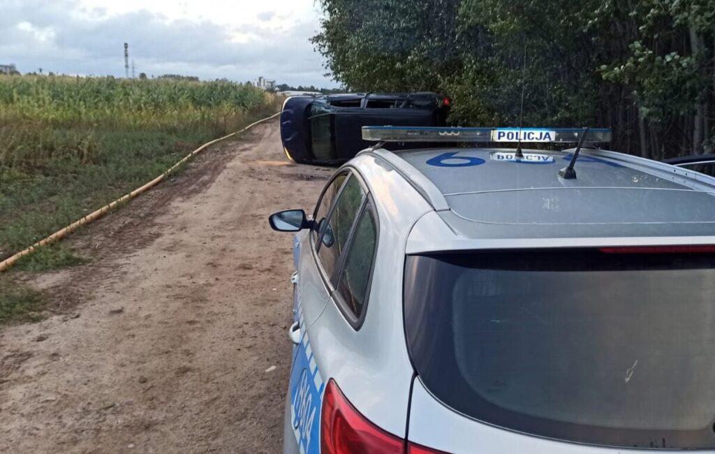Policyjny radiowóz zaparkowany na drodze, obok przewrócony samochód osobowy w otoczeniu pól.