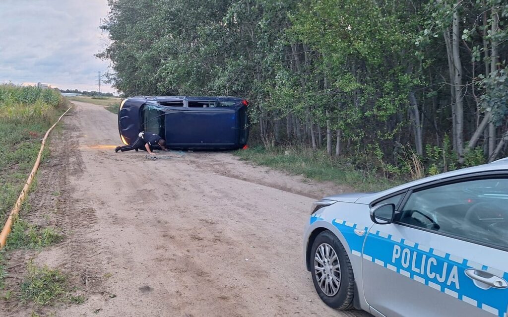 Mężczyzna w czarnym ubraniu przeszukuje przewrócone auto na poboczu drogi, obok stoi policyjny radiowóz.