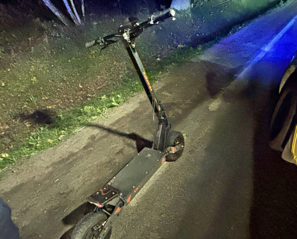 Uszkodzona elektryczna hulajnoga leży na jezdni obok zaparkowanego samochodu, oświetlona niebieskim światłem.