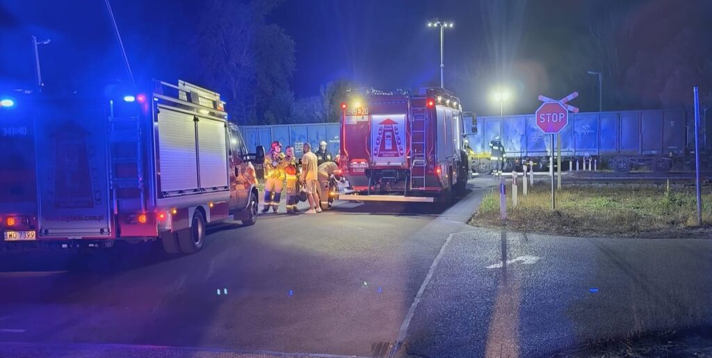 Grupa strażaków w mundurach omawia sytuację obok wozu strażackiego na drodze w pobliżu przejazdu kolejowego.