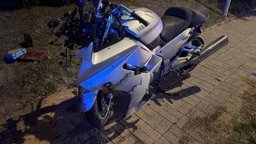 Zniszczony motocykl marki Yamaha stoi na chodniku, z uszkodzeniami karoserii i przodem. W tle widoczna trawa i fragmenty drogi.