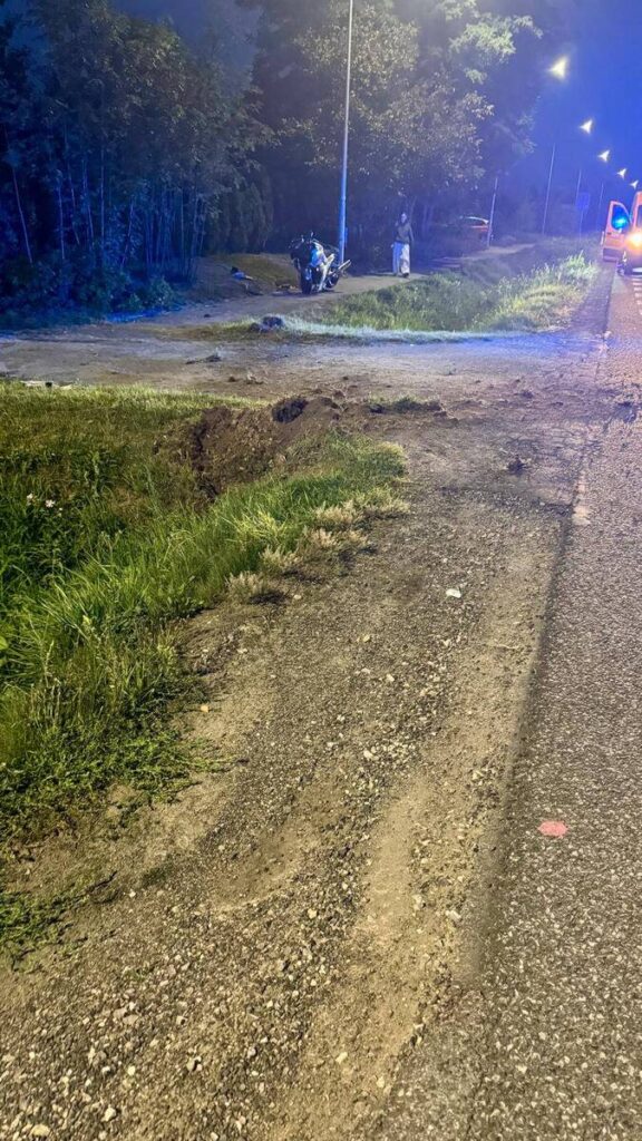 Motocyklista stoi obok uszkodzonego pojazdu na poboczu drogi, otoczony przez trawę i drzewa.