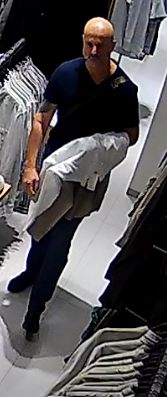 Mężczyzna w ciemnej koszulce i spodniach idzie między wieszakami z ubraniami w sklepie odzieżowym.
