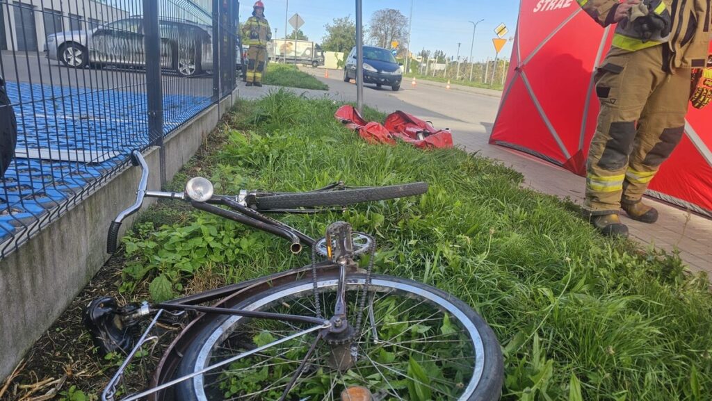 Rower leży na trawie obok drogi, a w tle widać strażaka w mundurze i czerwoną namiotową osłonę.