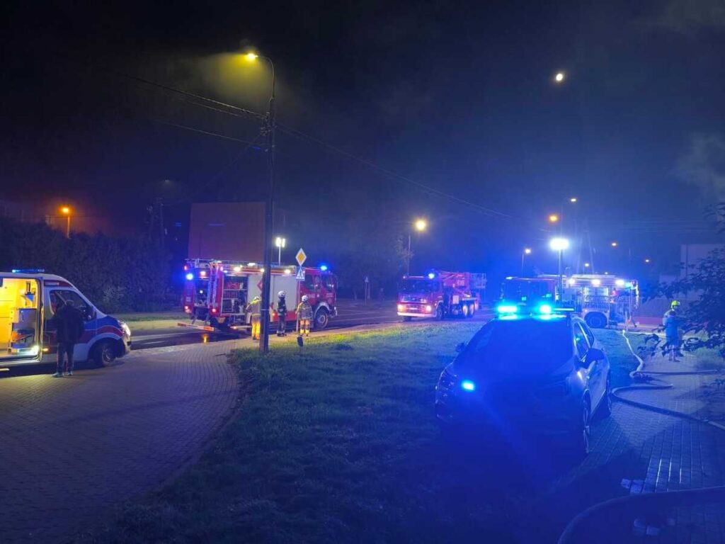 W nocy na ulicy pracują strażacy przy gaszeniu pożaru; w tle widoczne wozy strażackie i karetka.