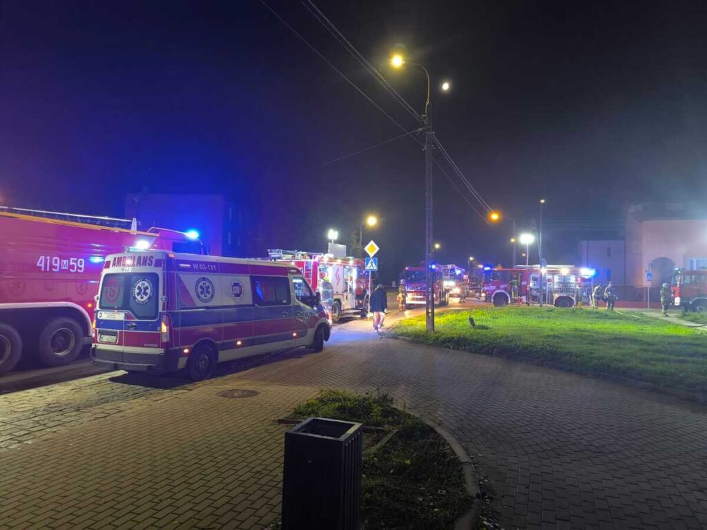 Na ulicy w nocy zaparkowane karetki i wozy strażackie, w tle widać oświetlony budynek.