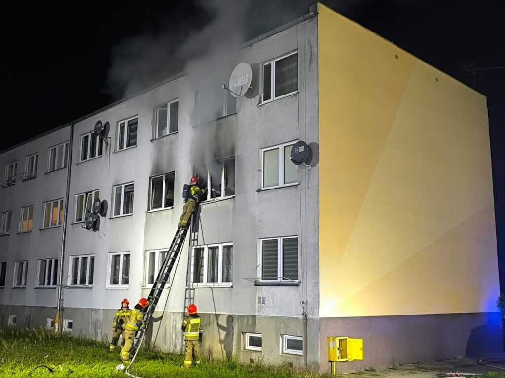Strażak na drabinie gaszący ogień wydobywający się z okna bloku mieszkalnego w nocy, obok inni strażacy przygotowujący sprzęt.