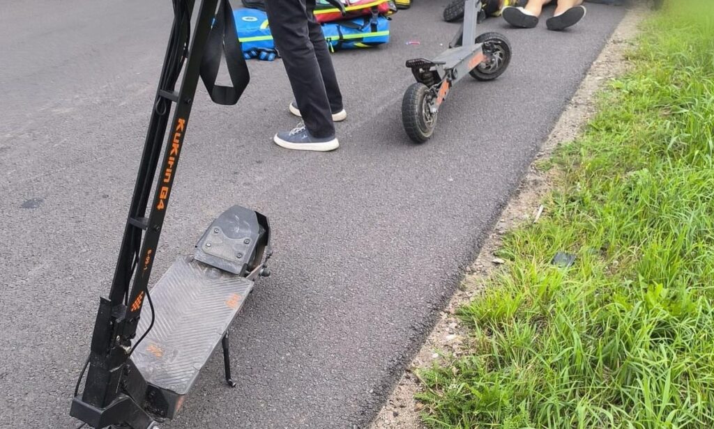 Na chodniku stoją elektryczne hulajnogi, obok nich osoby w casualowych ubraniach z plecakami.