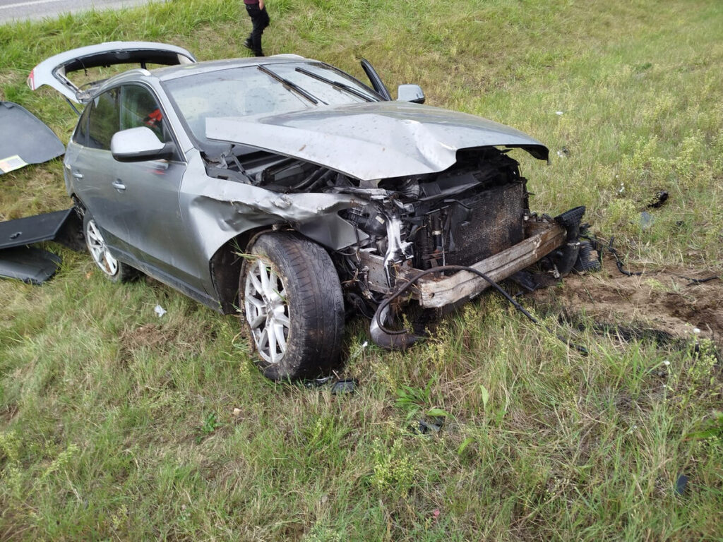 Uszkodzony samochód osobowy leży na poboczu drogi, z wgniecioną przednią częścią i otwartą maską. Obok widoczne resztki elementów pojazdu.