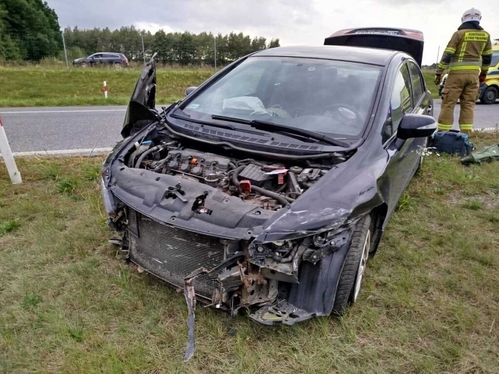 Uszkodzony samochód osobowy z otwartą maską stoi na poboczu drogi, obok widać strażaka w mundurze.