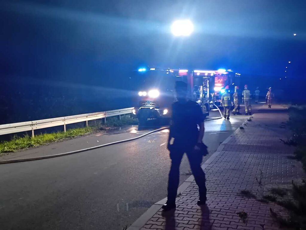 Strażacy w mundurach pracują przy wozie strażackim z włączonymi sygnałami świetlnymi na nocnej drodze.
