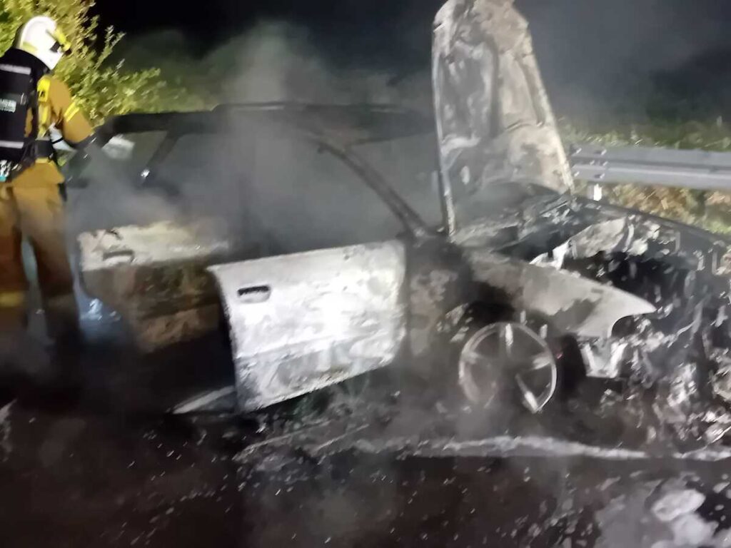 Strażak w specjalnym ubraniu gasi palący się wrak samochodu, otoczonego gęstym dymem.