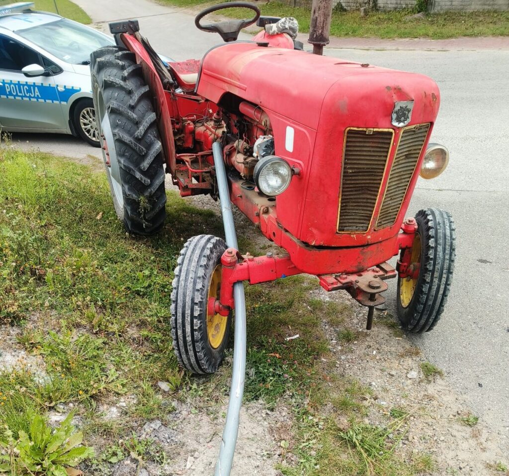 Czerwony traktor z metalowym wężem stoi na poboczu drogi obok policyjnego samochodu.