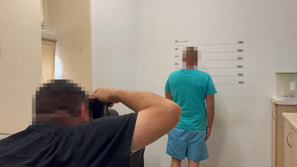 Mężczyzna w zielonej koszulce stoi tyłem przy ścianie z oznaczeniami wzrostu, fotografowany przez osobę z aparatem.