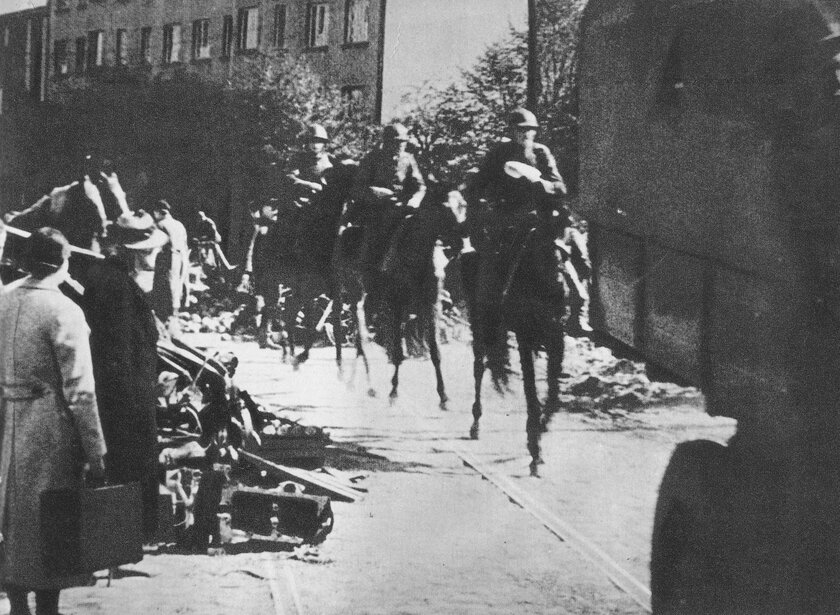 Grupa żołnierzy na koniach przejeżdża ulicą, obok stojący mężczyźni z walizkami i porzuconymi bagażami.