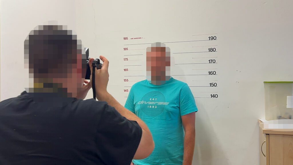 Mężczyzna w niebieskiej koszulce stoi na tle ściany z oznaczeniami wysokości, podczas gdy fotografuje go inna osoba.