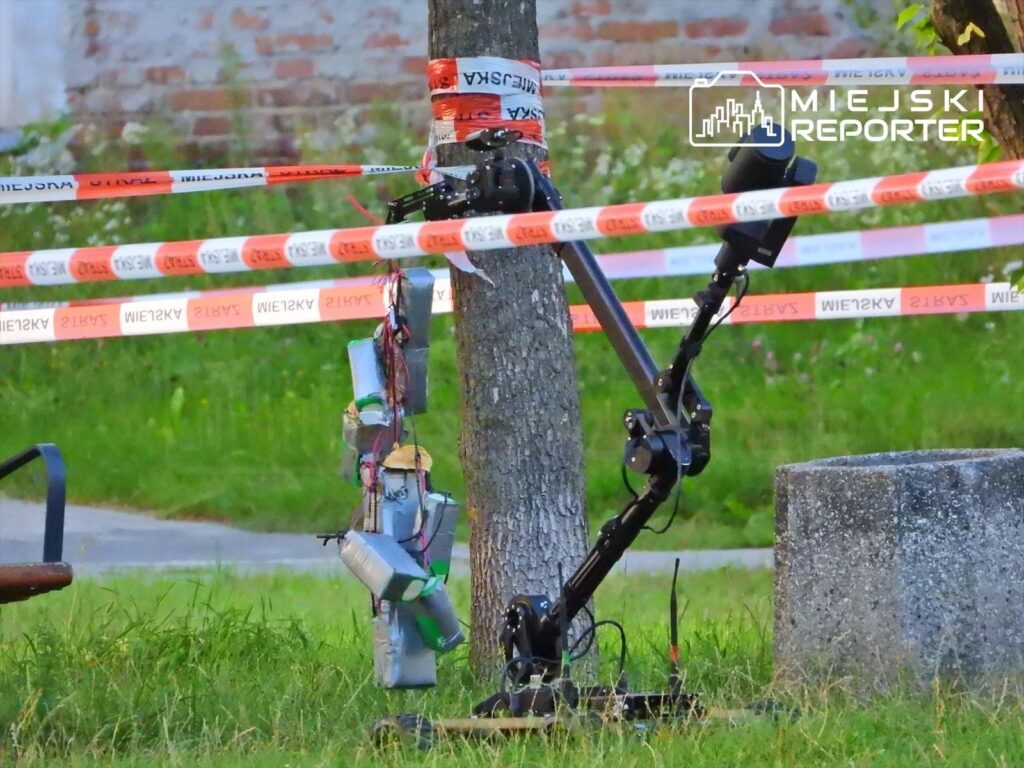 Robot z ramieniem mechanicznym zbliża się do zawieszonych przedmiotów na drzewie, otoczony taśmą policyjną.
