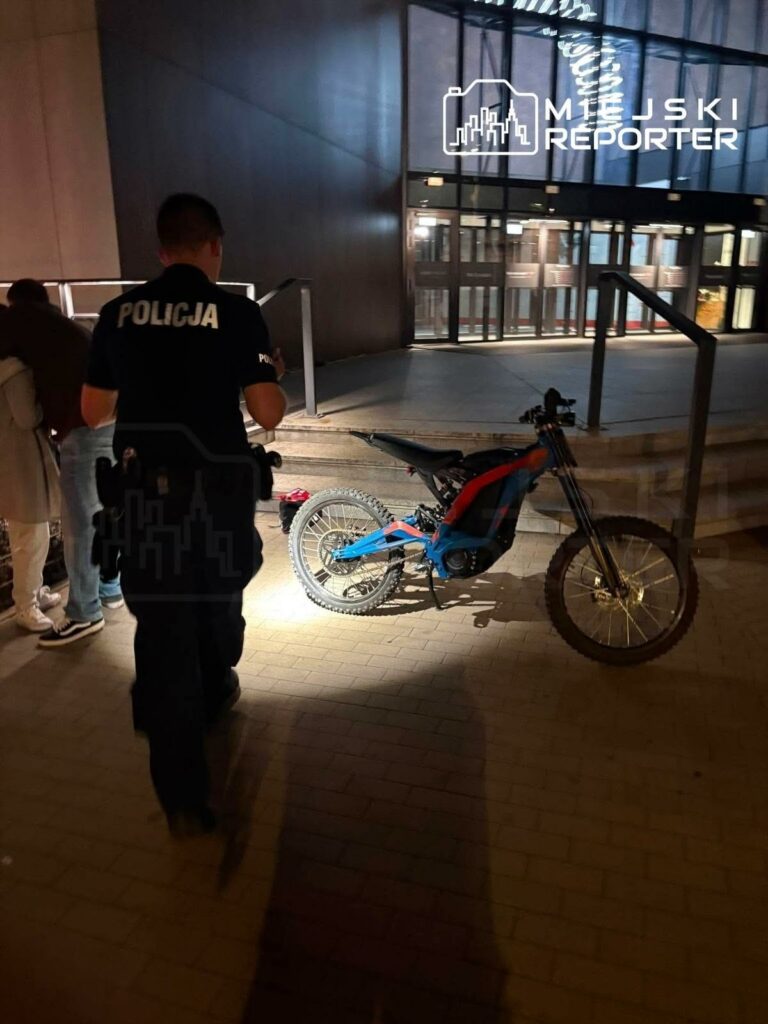 Policjant w mundurze stoi obok elektrycznego motocykla na chodniku przed nowoczesnym budynkiem.