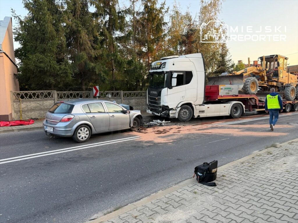 Zderzenie samochodu osobowego z ciężarówką na drodze, obok leży uszkodzony pojazd, a policjant w kamizelce podchodzi do wypadku.