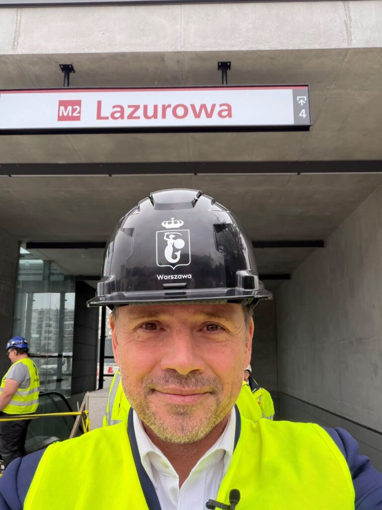 Mężczyzna w hełmie i kamizelce odblaskowej stoi przed oznakowaniem stacji metra M2 Lazurova.
